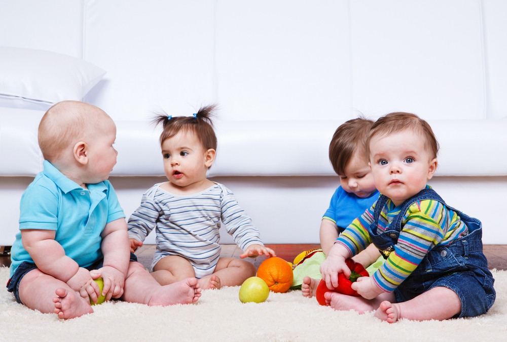 Four babies sitting on a soft carpet with colorful toys around them.