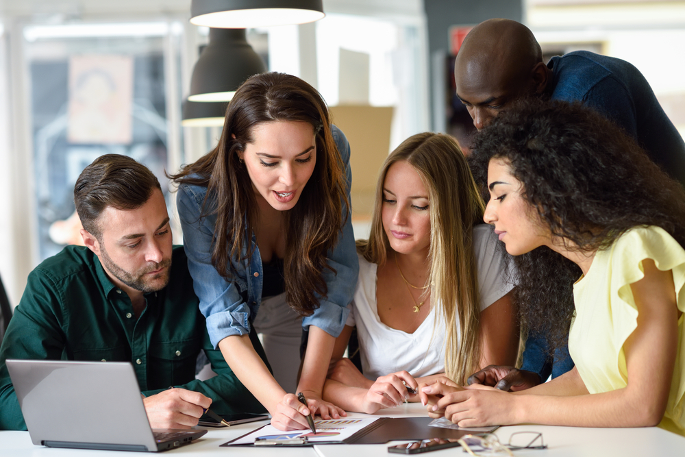 A team collaborates around a table, discussing a project with documents and digital devices.
